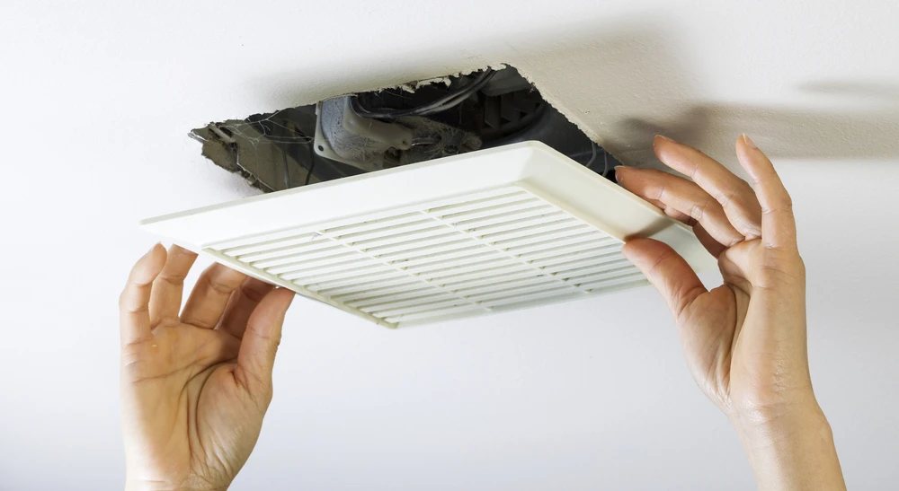 Mains installant une grille d'aération blanche dans un plafond résidentiel, accès visible au conduit de ventilation dans les combles