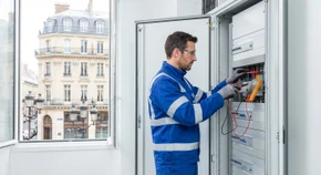 Électricien qualifié en tenue professionnelle effectuant une installation électrique ou de contrôle de conformité sur un tableau électrique ou une installation murale. L'image doit montrer clairement un professionnel du domaine électrique en action, de manière sérieuse et professionnelle, dans un environnement d'installation électrique réelle (domicile, bâtiment tertiaire ou industriel).