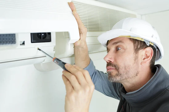 Un technicien en casque de s&eacute;curit&eacute; blanc installe ou r&eacute;pare un syst&egrave;me de ventilation au plafond d'un int&eacute;rieur moderne