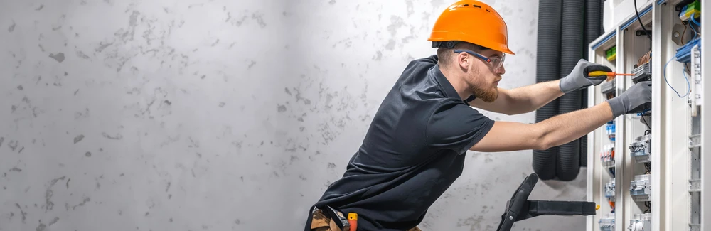 Électricien équipé d'un casque orange et de lunettes de protection travaille sur un tableau électrique industriel